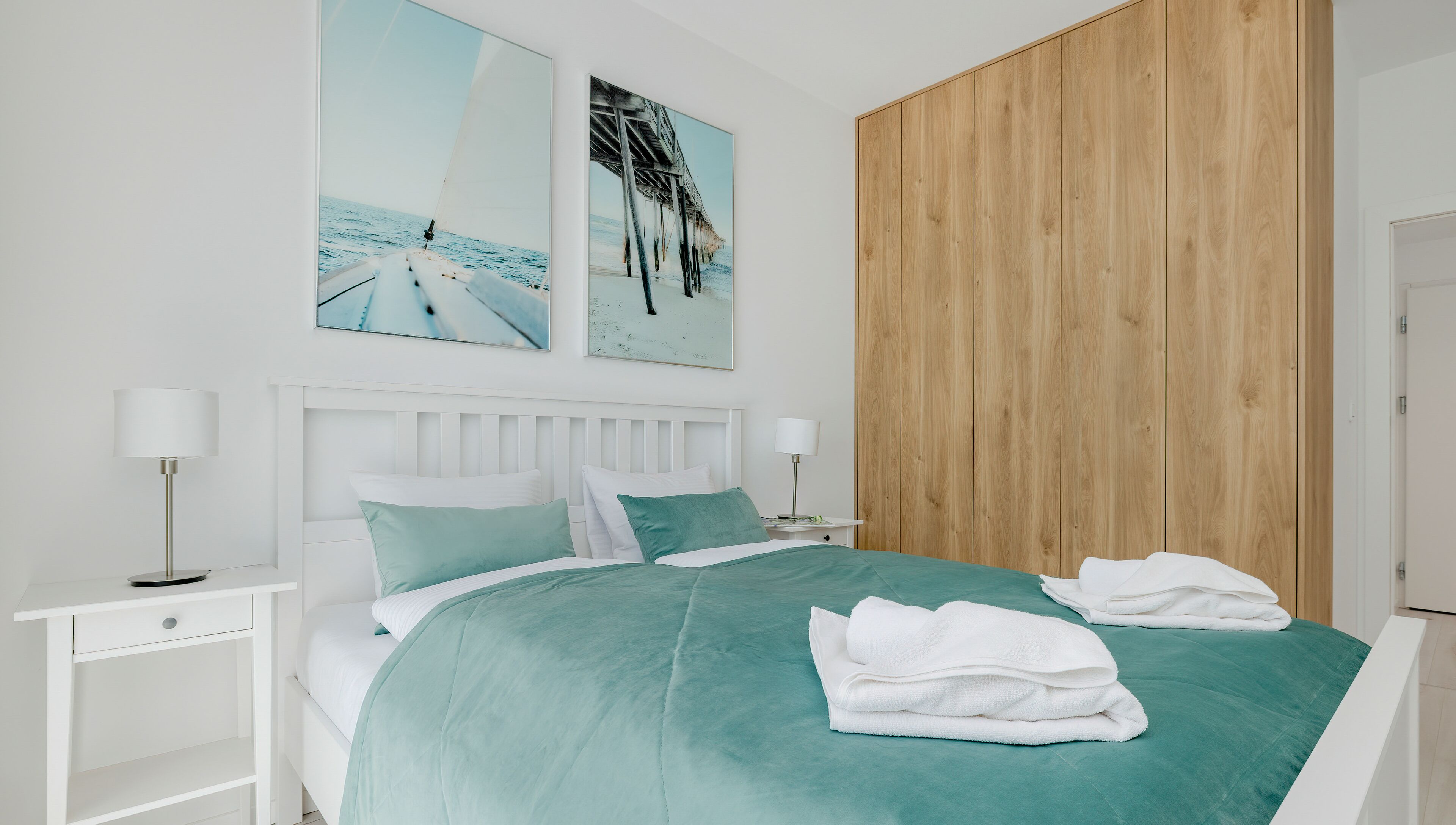 A modern bedroom with a bed, white nightstands, a wooden panel wall, and two beach-themed paintings.