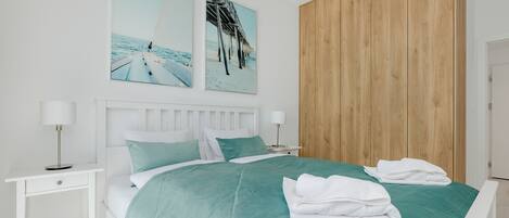 Un dormitorio moderno con cama, mesitas de noche blancas, un panel de pared de madera y dos cuadros con motivos playeros.