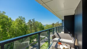 Een balkon met glazen hekwerk, rieten meubilair en uitzicht op bos en water.