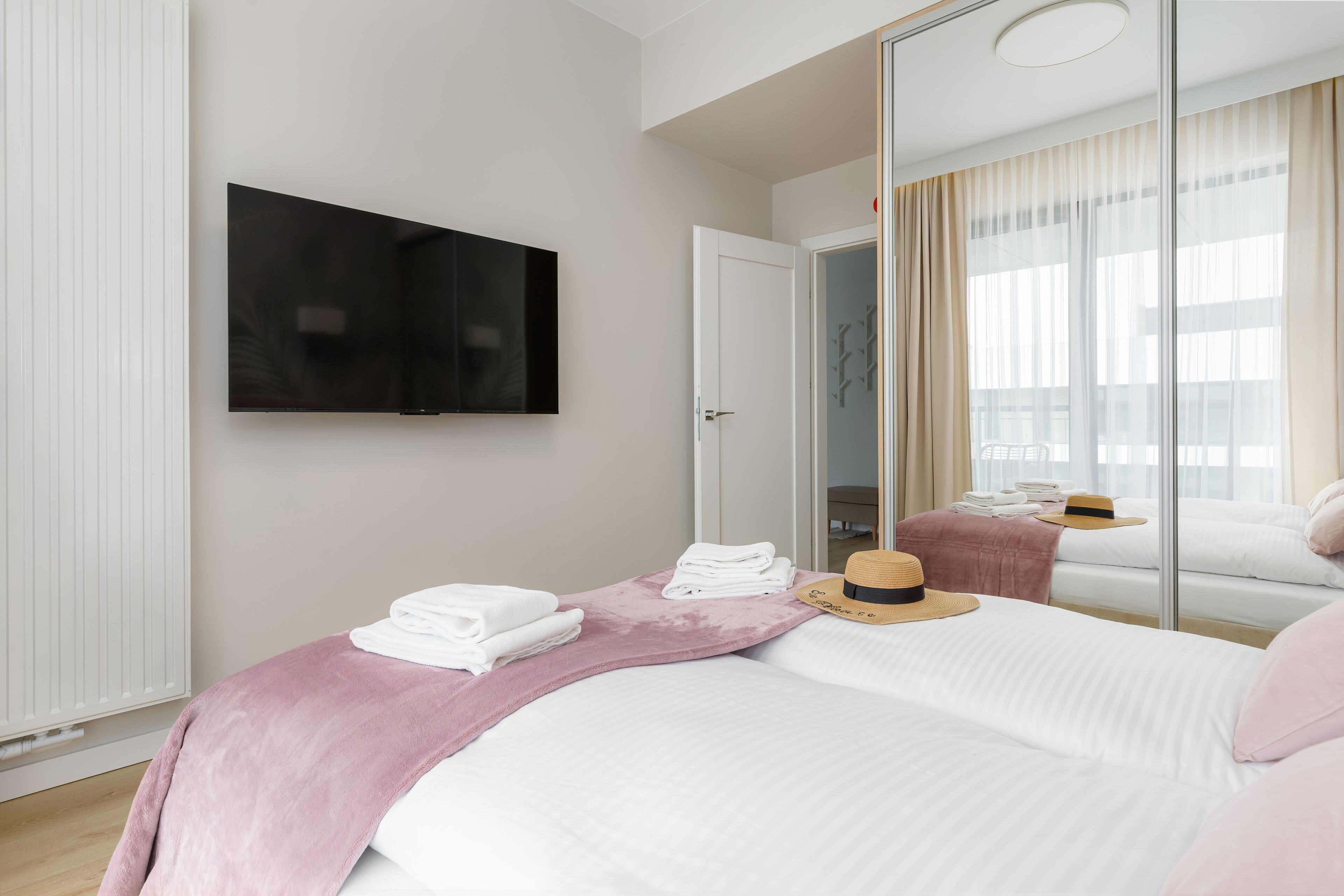 Ein Hotelzimmer mit einem Bett, einem wandmontierten Flachbildfernseher, einem Fenster mit transparenten Vorhängen und einem Spiegel, der den Raum widerspiegelt.