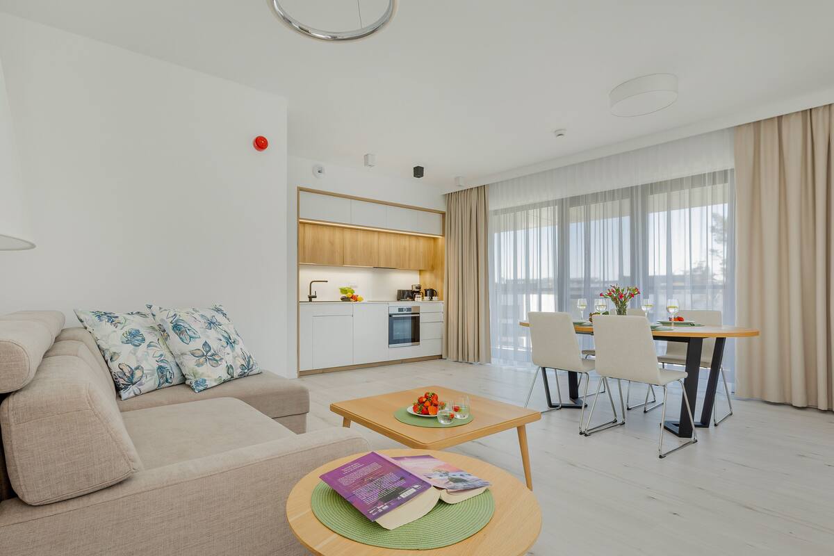 A modern living room with a sofa, a coffee table with books, a dining area with a table set for two, and a kitchen in the background.