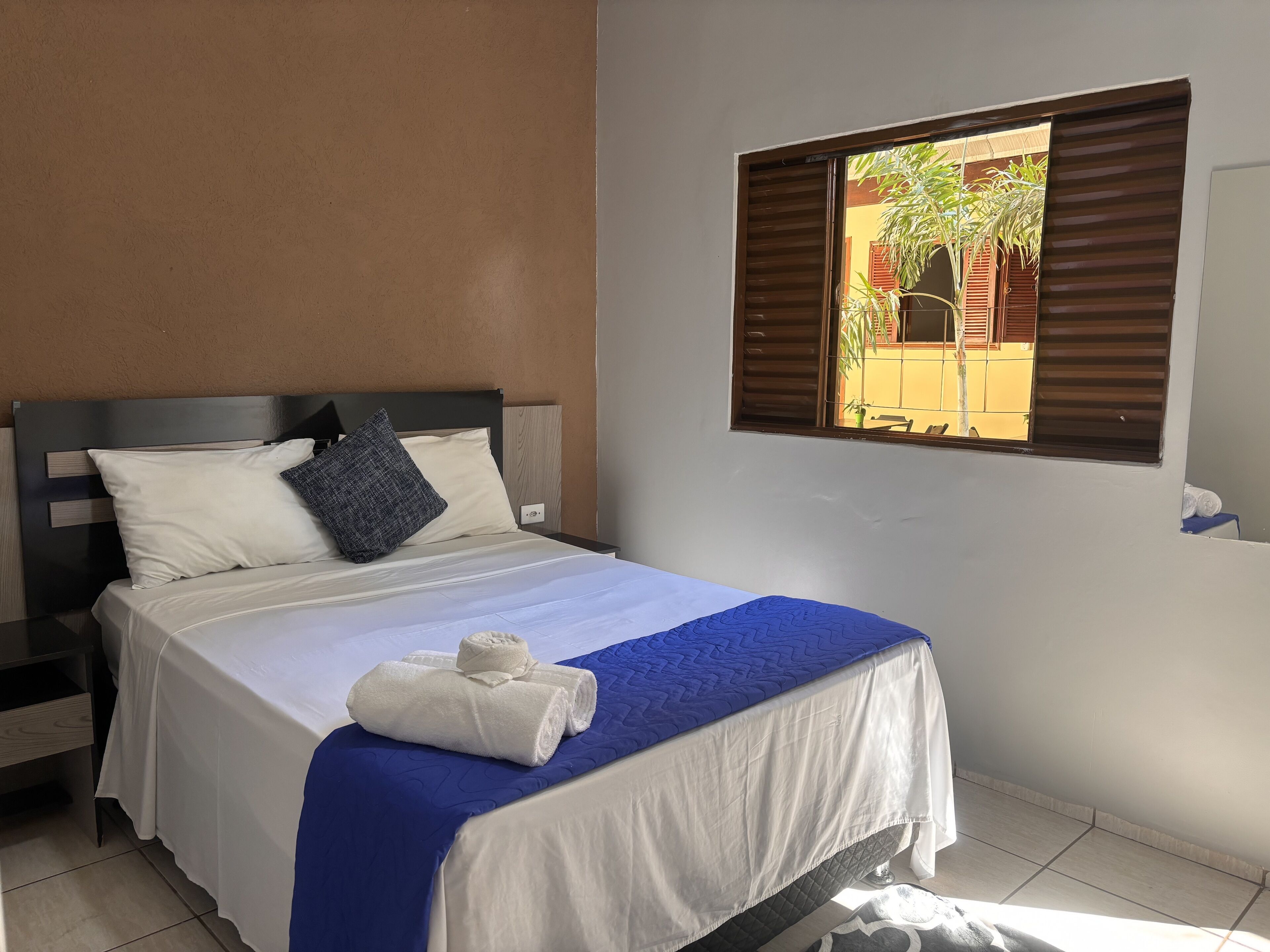 Cama bem arrumada com roupa de cama branca e azul, cabeceira de madeira e janela com persianas.