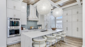 Una cocina moderna con armarios blancos, una isla central con taburetes de bar, electrodomésticos de acero inoxidable e iluminación colgante hexagonal.