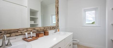 A bathroom with white tiles, a stone accent wall, a large mirror, and a window.