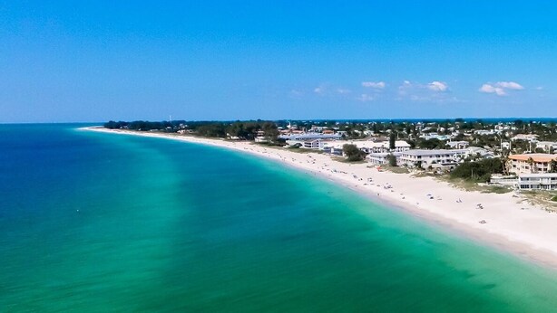 An aerial view of a coastal area with a long sandy beach, clear turquoise waters, and a row of buildings along the shore. - Beachfront Bliss with Gulf Views & Heated Pool (Holmes Beach)