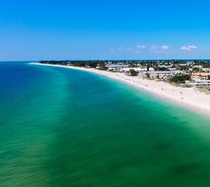 An aerial view of a coastal area with a long sandy beach, clear turquoise waters, and a row of buildings along the shore. - Beachfront Bliss with Gulf Views & Heated Pool (Holmes Beach)