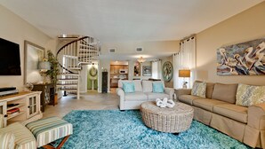 A spacious living room with a large sofa, a round wicker ottoman, a striped armchair, and a TV on a cabinet. A spiral staircase leads to an upper level. - Beachfront Bliss with Gulf Views & Heated Pool (Holmes Beach)