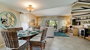 A spacious living area with a dining table set for six, surrounded by wicker chairs. A large decorative mirror and a chandelier are visible. The room features a neutral color palette and a view of the outdoors through large windows. - Beachfront Bliss with Gulf Views & Heated Pool (Holmes Beach)