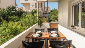 A balcony with a wooden table set for dining, surrounded by lush greenery and a view of neighboring buildings.