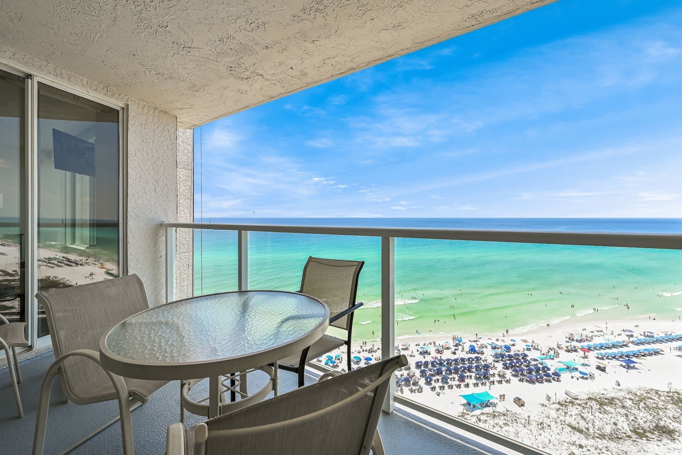 Een balkon met een glazen tafel en stoelen, uitkijkend op een strand met parasols en mensen.
