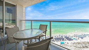 Ein Balkon mit Glastisch und Stühlen, der Blick auf einen Strand mit Sonnenschirmen und Personen freigibt.