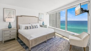 Ein Schlafzimmer mit großem Fenster, einem Bett, einem Nachttisch, einem Sessel und einem an der Wand montierten Fernseher.
