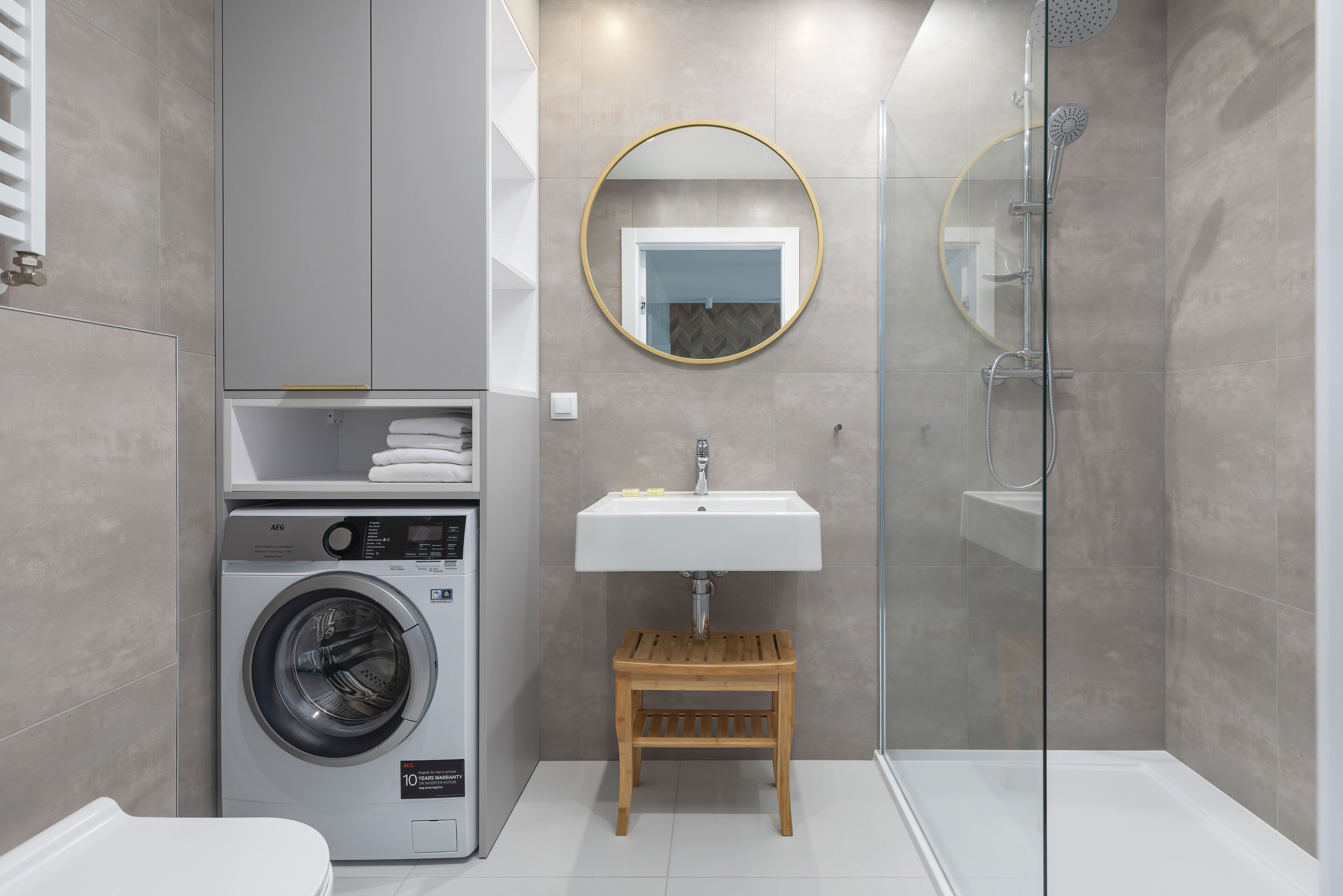A modern bathroom with a washing machine, a sink, a wooden stool, and a glass-enclosed shower.