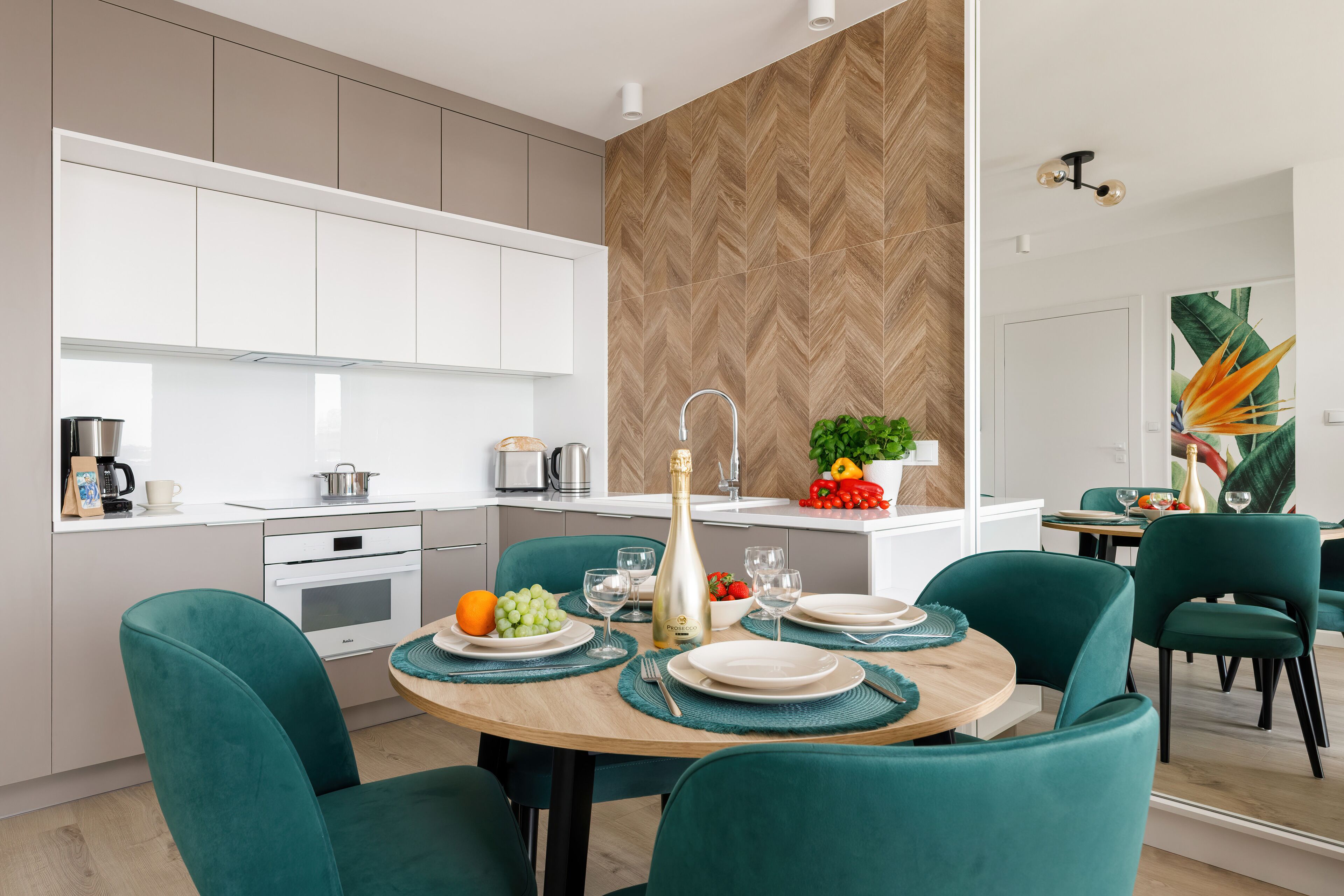 Una cocina moderna con armarios blancos, un salpicadero de madera en espiga y un área de comedor con sillas color turquesa y una mesa de madera para dos personas.