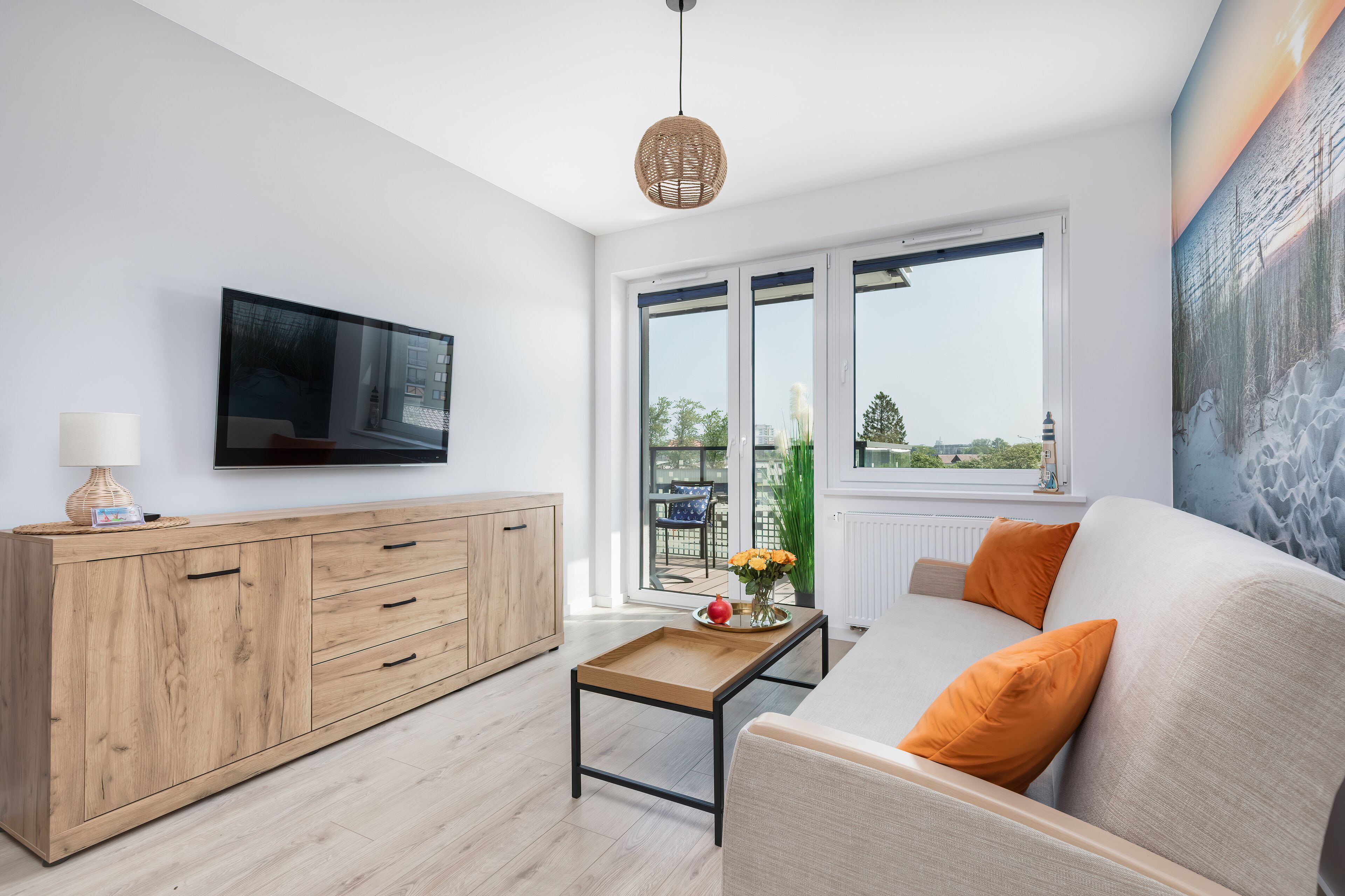 A modern living room with a wooden cabinet, a sofa with orange cushions, a coffee table with flowers, and a large window offering a view of the outdoors.