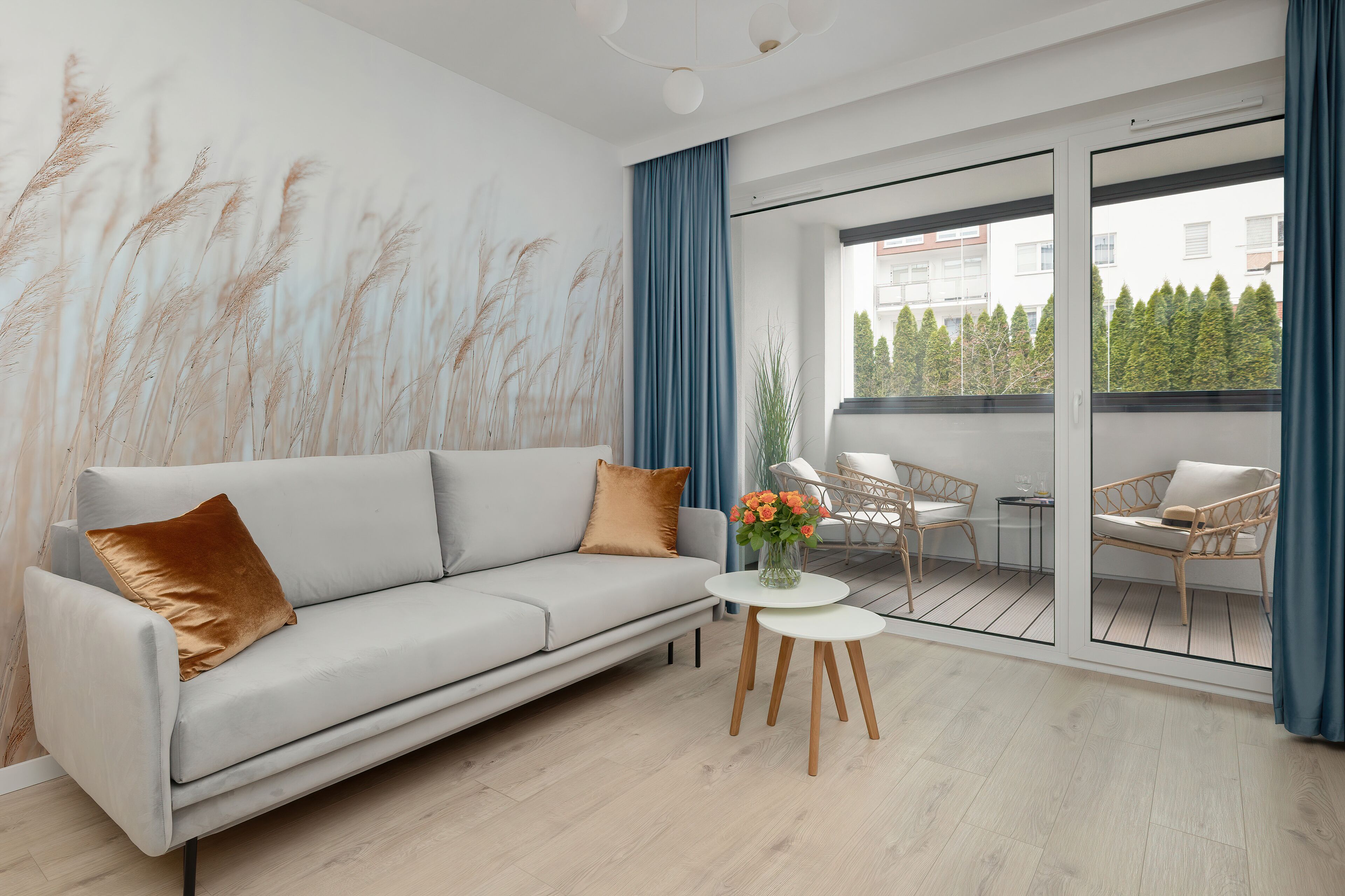 A modern living room with a sofa, a small round table with a vase of flowers, and a view of a balcony with outdoor furniture.