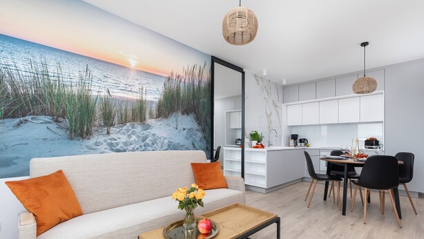 Una sala moderna con sofá, una mesita de centro y un comedor. Se observa una cocina al fondo con armarios blancos y una mesa de comedor.