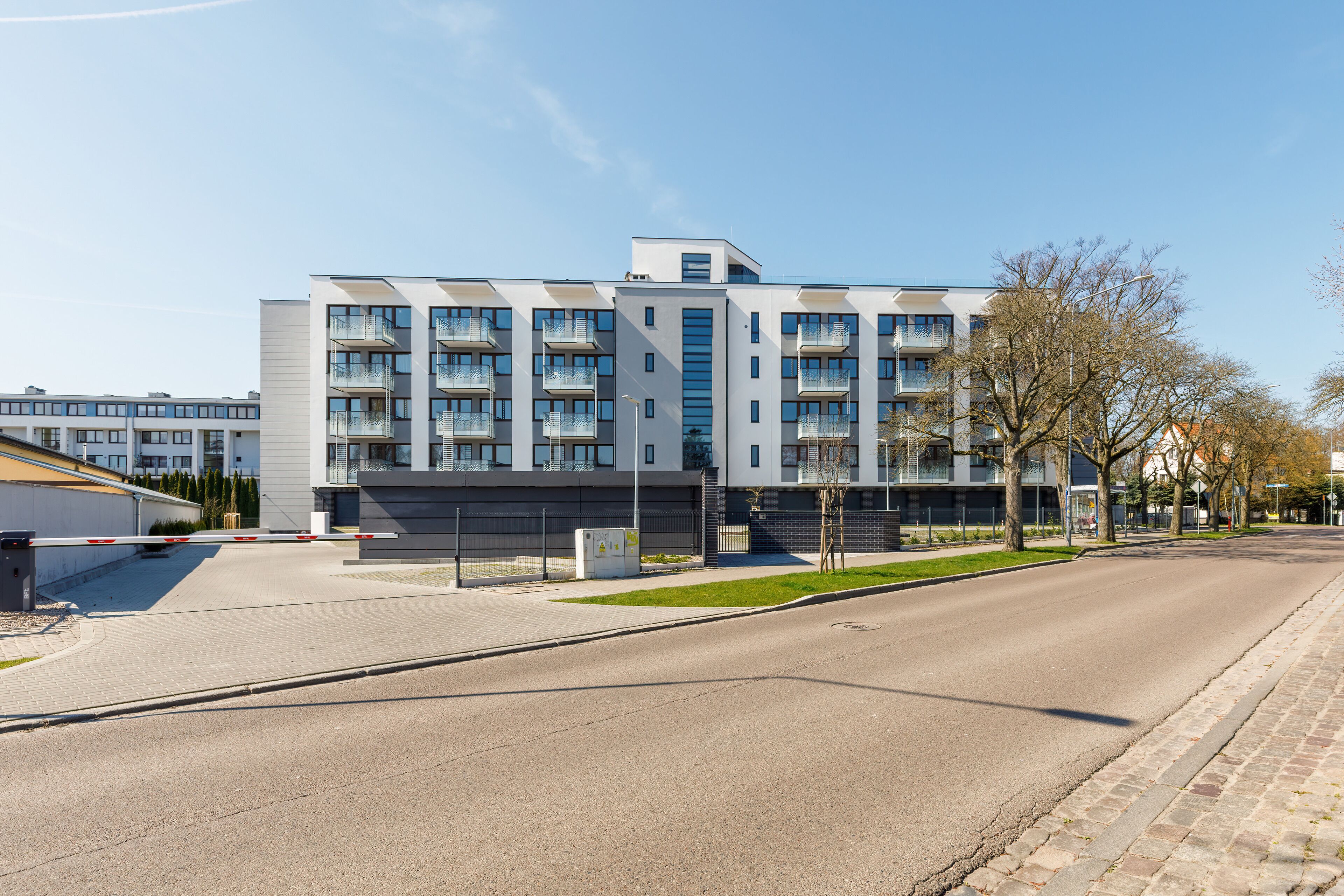 A modern multi-story building with balconies, a gated entrance, and a paved road in front.