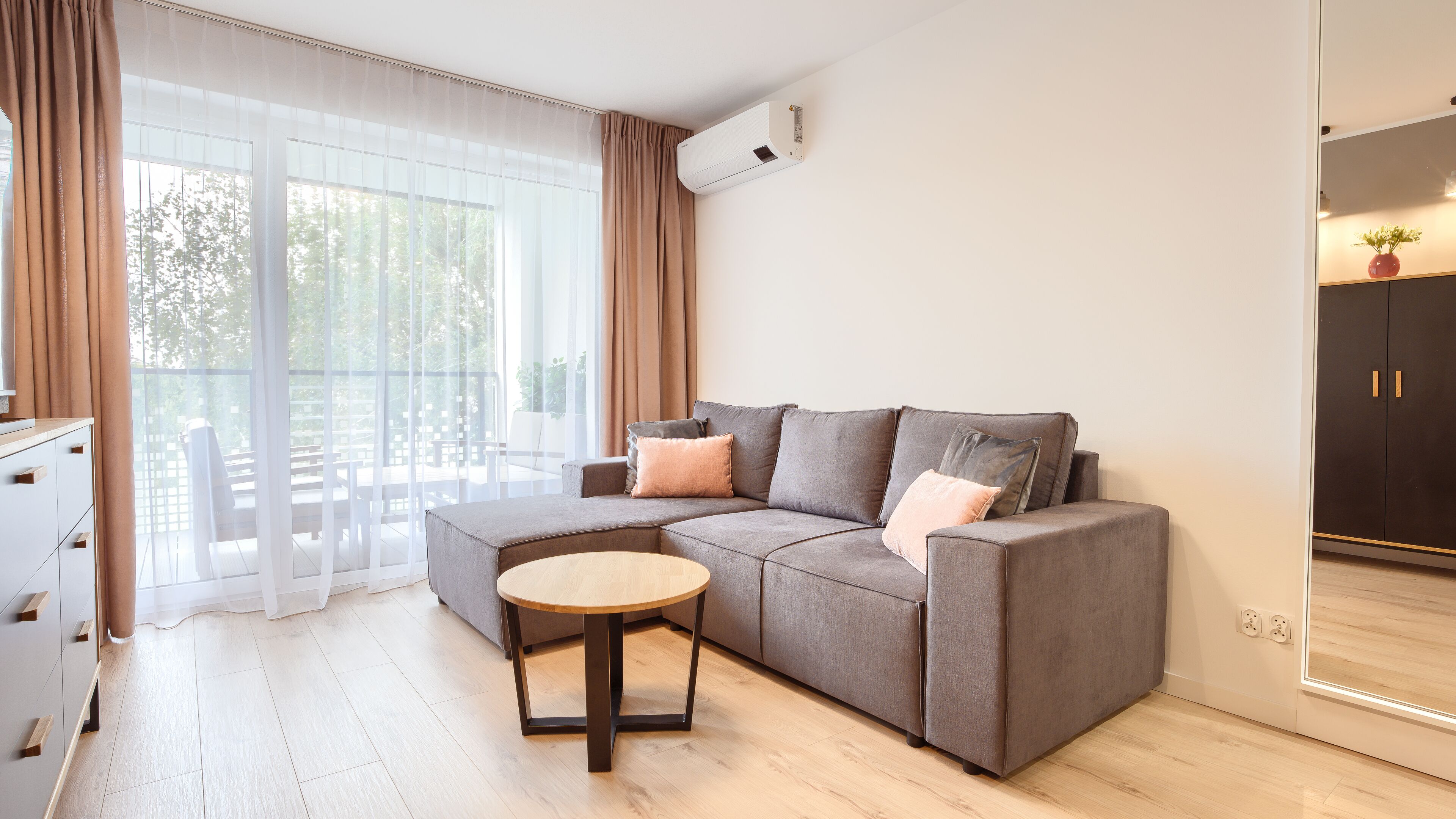 Een moderne woonkamer met een grijze modulaire bank, een ronde houten salontafel en een schuifdeur naar een buitenruimte.
