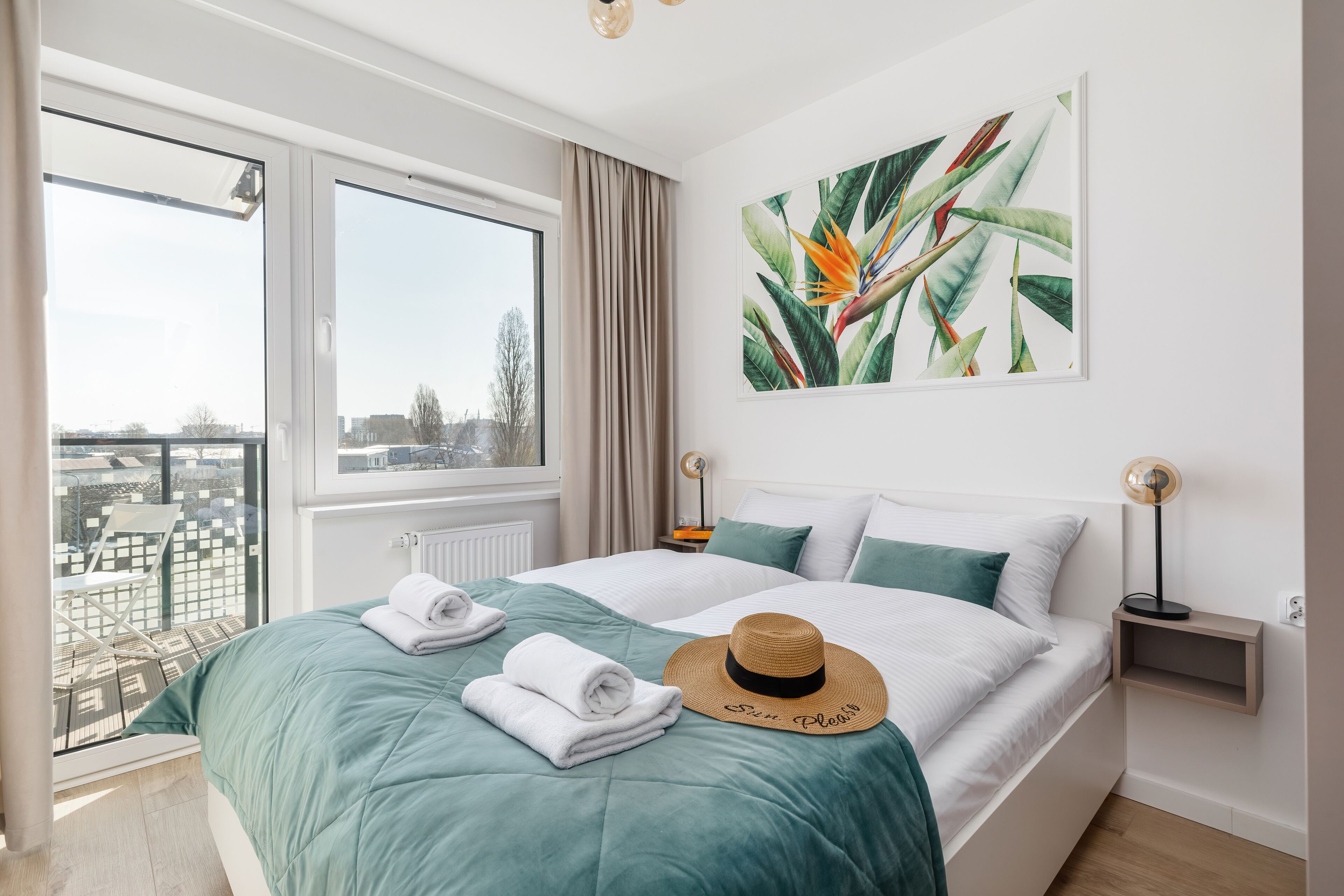 A bedroom with a large bed, a view of the city, a tropical plant painting, and a balcony with a chair.