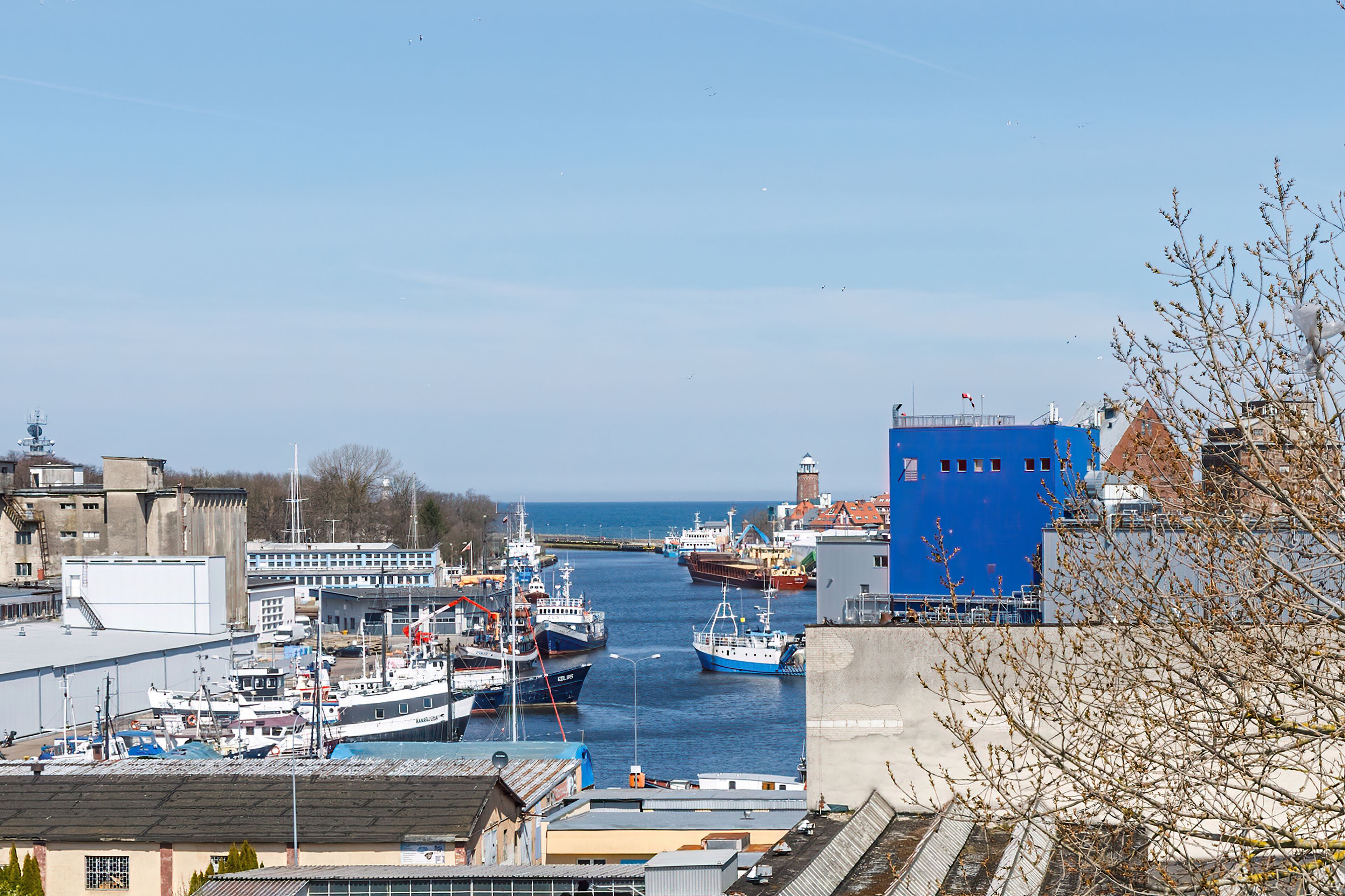 A harbor with boats docked, a blue building, and a lighthouse in the distance.