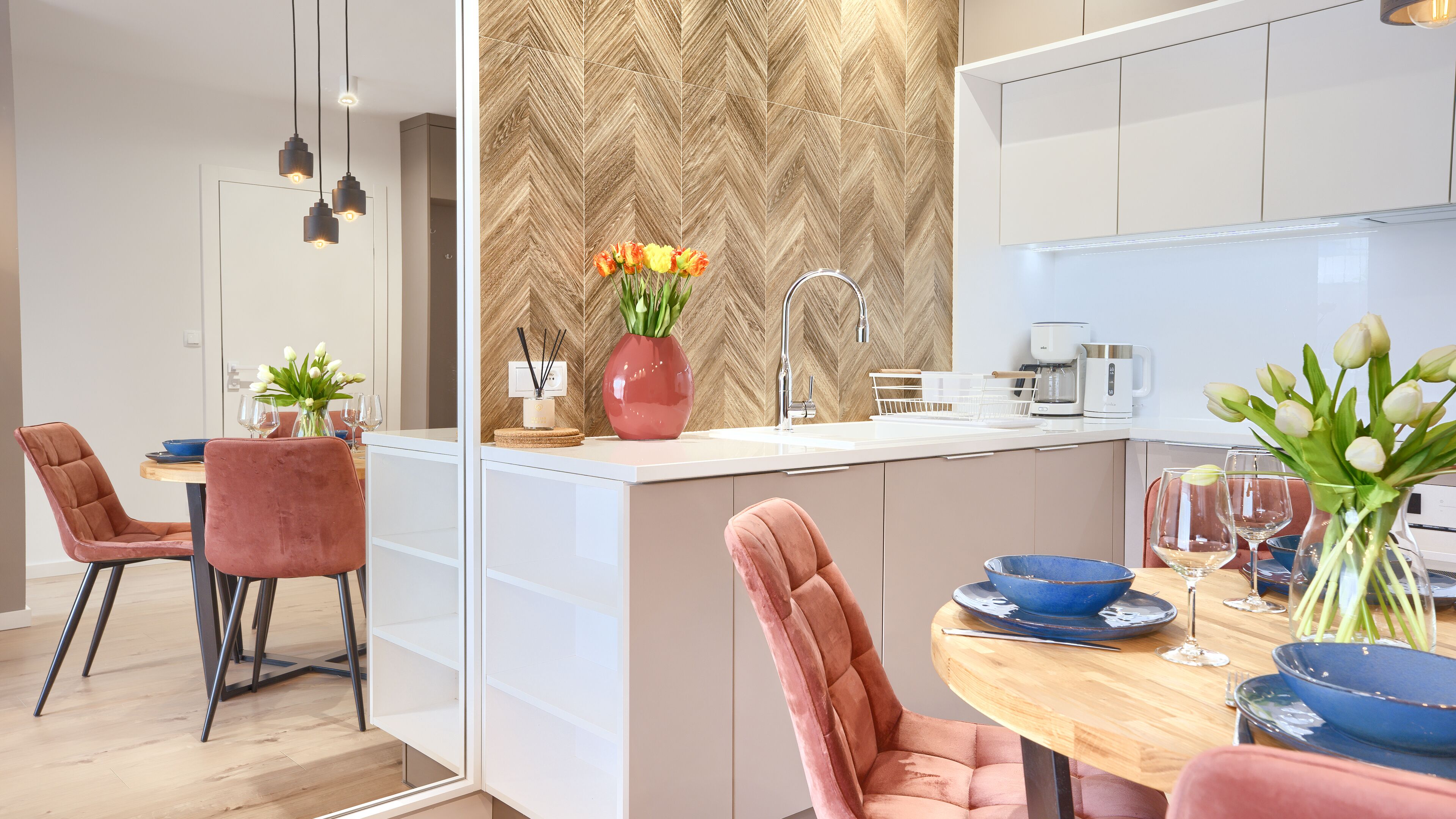 Una cucina moderna con zoccolo a spina di pesce, un'isola centrale con lavello e un'area pranzo con sedie rosa e tavolo in legno.