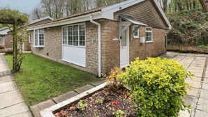 Une petite maison en briques avec une porte et des fenêtres blanches, entourée d’un jardin et d’arbres.