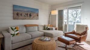 Un salón con un sofá modular, un sillón de cuero, un otoman tejido y un cuadro de una manada de caballos.