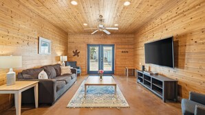 A cozy cabin interior with a wooden interior, a ceiling fan, a large TV, and a sofa. - Bron's Beach Shack with Pool & Near Beach (Port Aransas)