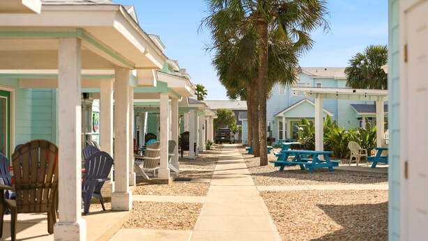 A covered outdoor area with picnic tables and chairs, a pathway, and a row of buildings with a light blue exterior. - Bron's Beach Shack with Pool & Near Beach (Port Aransas)