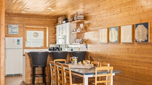 A cozy kitchen with wooden walls, a white refrigerator, a dining table with chairs, and framed artwork on the wall. - Bron's Beach Shack with Pool & Near Beach (Port Aransas)