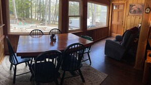 A wooden cabin with a dining table, chairs, and a view of the forest through large windows. - Kettle Cove in Sebago Lake, Maine (Casco)