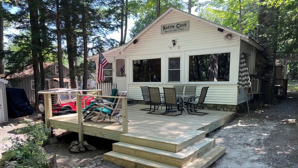 A small building labeled "Kettle Cove Bank and Post Office" with a deck, outdoor seating, and a flag. - Kettle Cove in Sebago Lake, Maine (Casco)