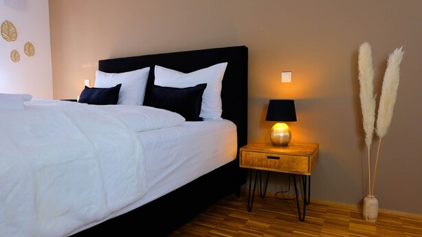 Ein ordentlich bezogenes Bett mit weißer und schwarzer Bettwäsche, ein Nachttisch mit einer Lampe und Pampasgras in einer Vase.