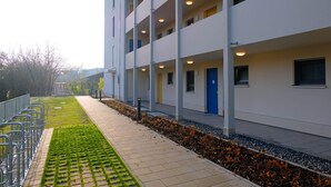 Ein modernes Gebäude mit mehreren Etagen, einem gepflasterten Weg und einer Grünfläche mit einem Metallgeländer an einer Seite.