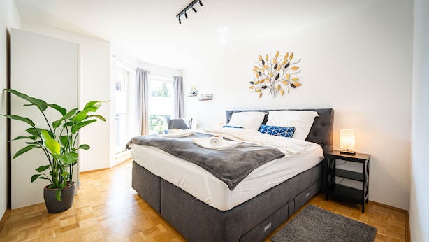 A modern bedroom with a grey bed, white bedding, a potted plant, a bedside table with a lamp, and a decorative wall piece. - Zentral Gelegenes 55 m² Design-apartment (Magdeburg)