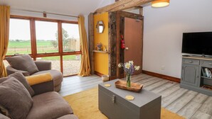 A living room with a sofa, a coffee table with decorative items, a television, and a view of a green landscape through large windows. - Bee Happy Barn (Norwich)