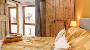 A bedroom with a bed, a wooden wardrobe, a window with curtains, and a lamp. - Bee Happy Barn (Norwich)