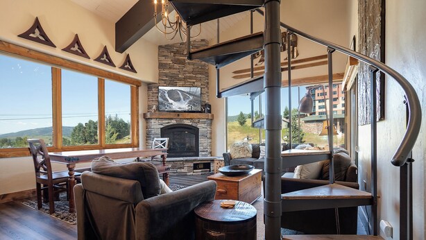A cozy living room with a stone fireplace, a curved staircase, and large windows offering a view of the outdoors.