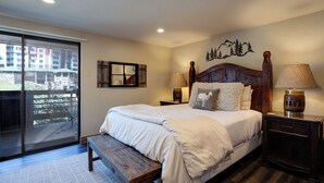 A bedroom with a large bed, wooden headboard, two bedside tables with lamps, a bench, a large window with a view of buildings, and a wall-mounted mirror.