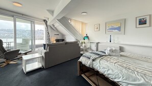 Une pièce moderne et bien éclairée, comprenant un lit, un canapé et une petite table. Un escalier mène à une mezzanine offrant une vue sur le paysage urbain.