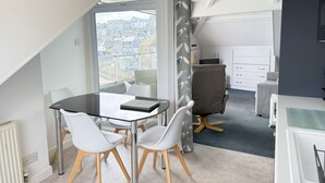 Une pièce moderne avec une table en verre, des chaises blanches, un fauteuil de bureau gris et une vue sur les immeubles à travers une grande fenêtre.