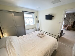 A bedroom with a bed, a window with blinds, a wall-mounted TV, and a stone fireplace. - Penryn a modern spacious ground floor apartment (St Ives)