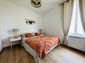 A bedroom with a wooden floor, a bed with a patterned blanket, two bedside tables with lamps, a wall-mounted decoration, and a window with curtains.