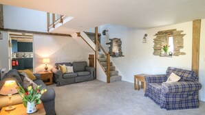 A cozy living room with a grey sofa, a blue plaid sofa, a wooden coffee table, and a small side table with a lamp and a vase of flowers. A staircase with wooden railings leads to an upper level.