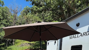 En campingplads med et picnicbord og en parasol, en hvid campingvogn med 'MALLARD' påtrykt, og et rødt køretøj i baggrunden.