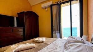 A hotel room with a bed, a wooden dresser, a window with a view of the sea, and an air conditioning unit. - SUNSET (Cefalù)