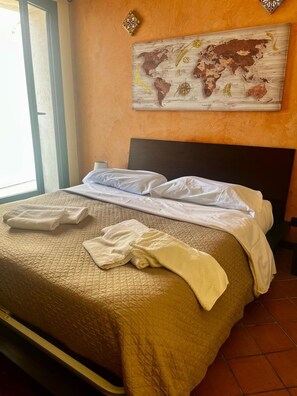Een netjes opgemaakt bed met witte lakens, een geborduurde beige deken en opgerolde handdoeken op het hoofdeinde. Een wereldkaart als kunstwerk hangt aan de muur boven het bed.