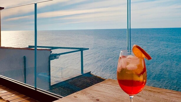 A glass of red drink on a wooden table with a view of the sea. - SUNSET (Cefalù)