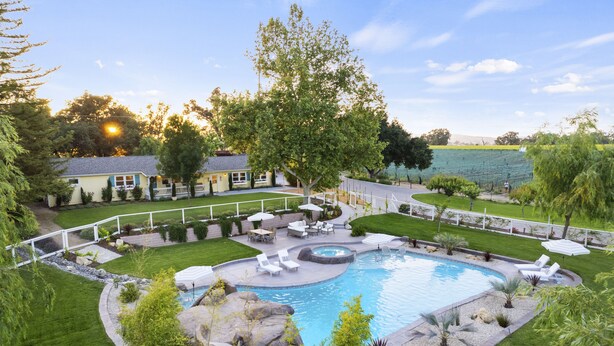 A backyard with a swimming pool, surrounded by a well-maintained lawn, a patio area with outdoor furniture, and a house in the background.